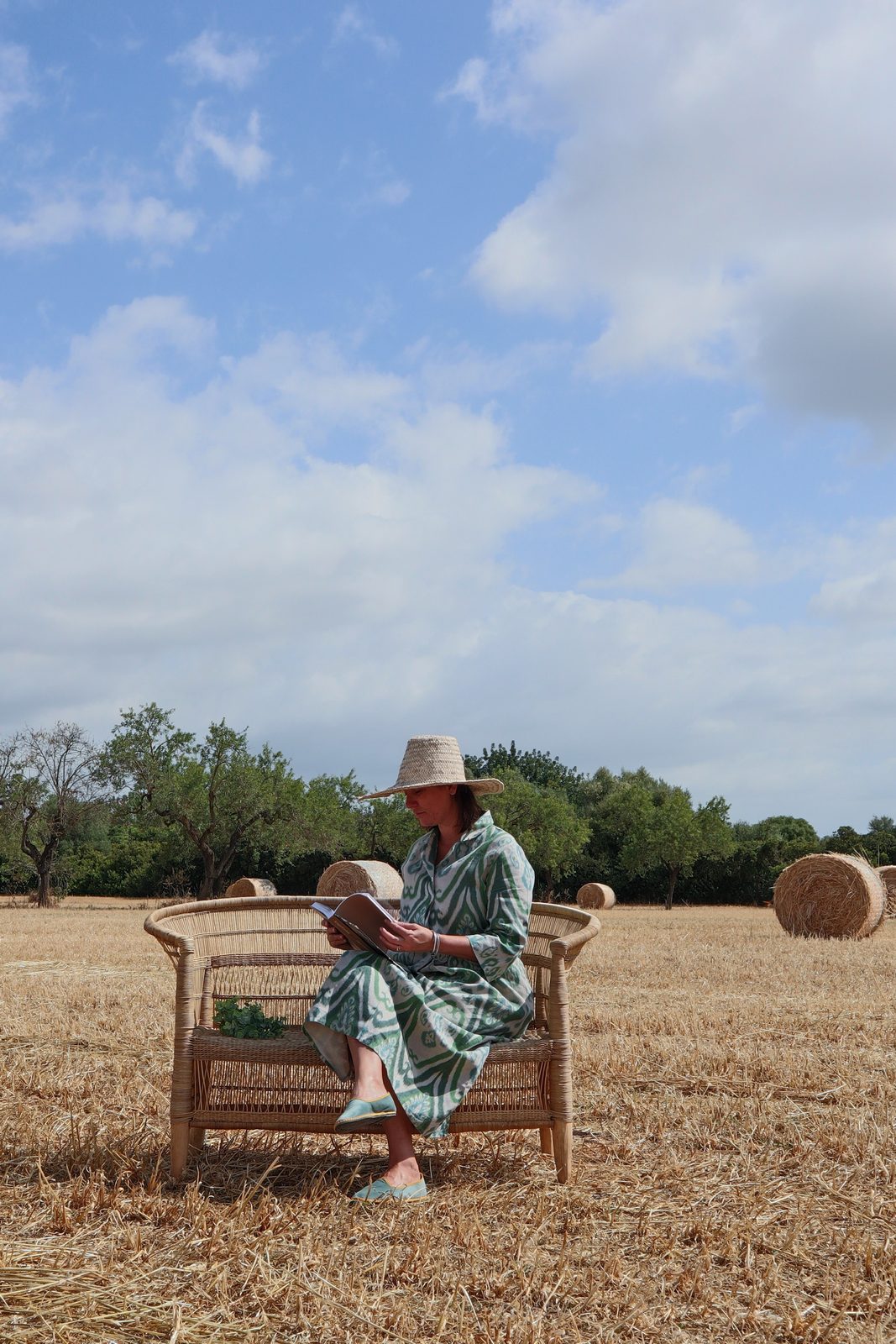 Woman in a striped dress and straw hat reading on a cane bench in a sunlit hay field